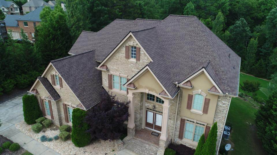 Arial photo of two story home with a third story loft area in the main section of the home. The multi level roof structure sports a series of pitch dormers inset within one another and a pyramid main roof for the central portion of the home. The house has brown asphalt tiles with a red accent color. The home facade is brown painted stucco and light stone