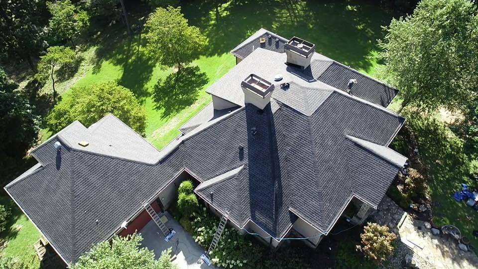 aerial photo of roof from almost dirctly above. The roofline shows a very high pitch roof with flat area at the peak. The roofline is pyramid shaped as it descends to dormers that surround the four sides of the main home. An enclosed breezeway leads to a multi-car garage that has the same pyramid roofline with a steeper pitch from the top as it does not have the flat area of the main building.