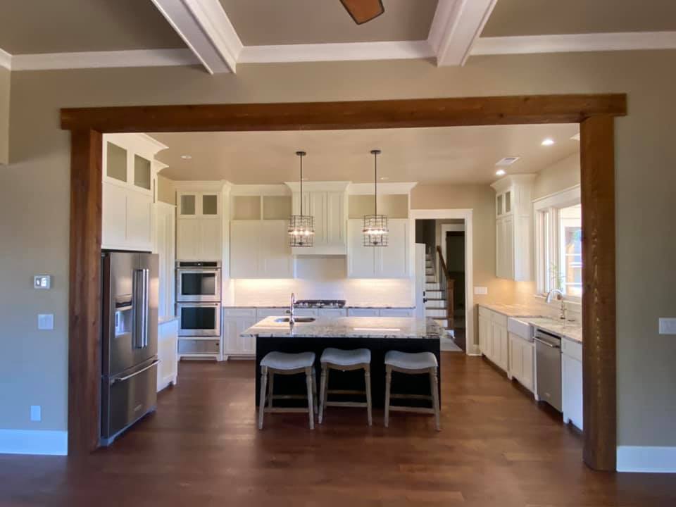 photo of chef's kitchen in the Boone House. It shows the entire wet bar kitchen Island with seating for three at the granite countertop, on the right the farm sink with large windows, the rear wall with range and ovens surrounded by floor to ceiling cabinets and on the left the french door refrigerator. Pendant lighting and wood floors.