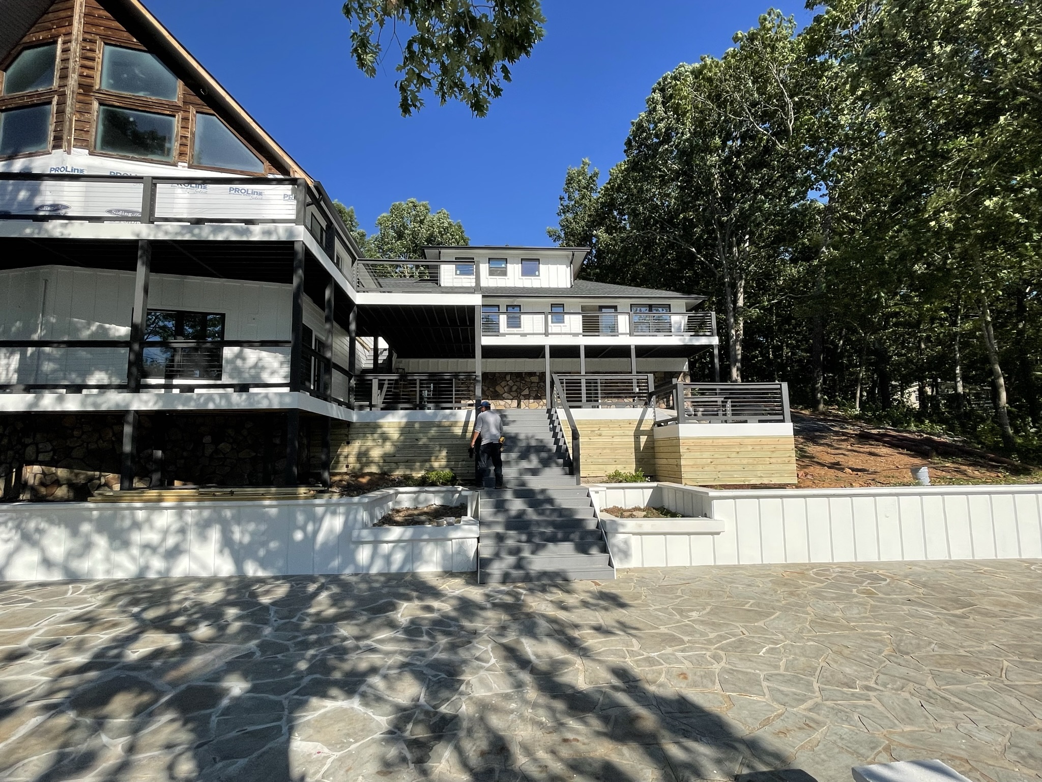 Wide view of house from the lake looking toward the rear of the home with stone patio, new terraces and decking system with paliedium window at great room still under construction